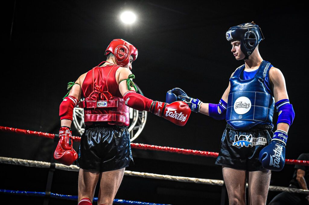 Two Muay Thai fighters in headgear and chest protectors touching gloves in the ring